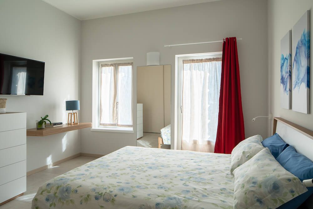 camera da letto con tende rosse, tv, cassettiera, specchio, mensola con elementi decorativi, letto con copriletto fiorato